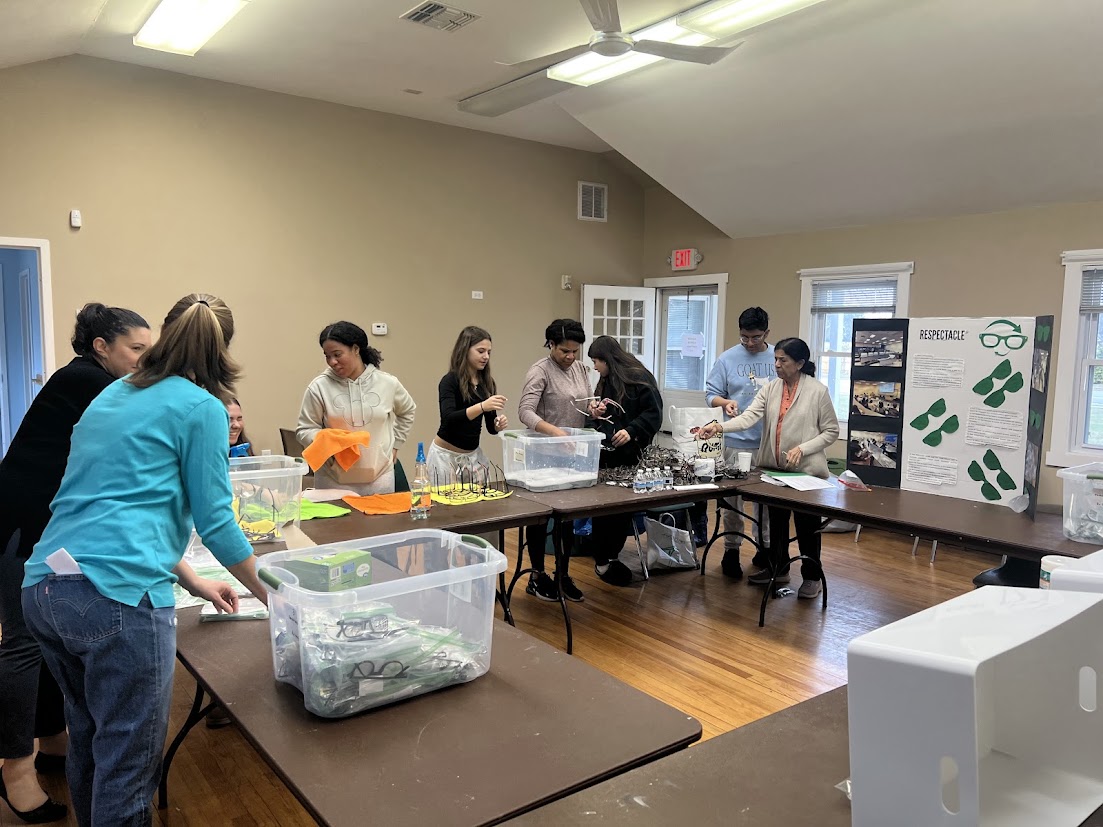 Volunteers cleaning and sorting donated eyeglasses at long tables beside a ReSpectacle display board